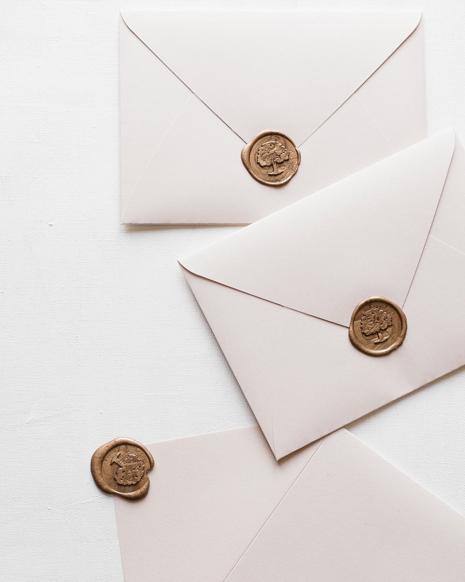 Three blush wedding envelopes sealed with gold wax stamps, arranged on a textured ivory backdrop